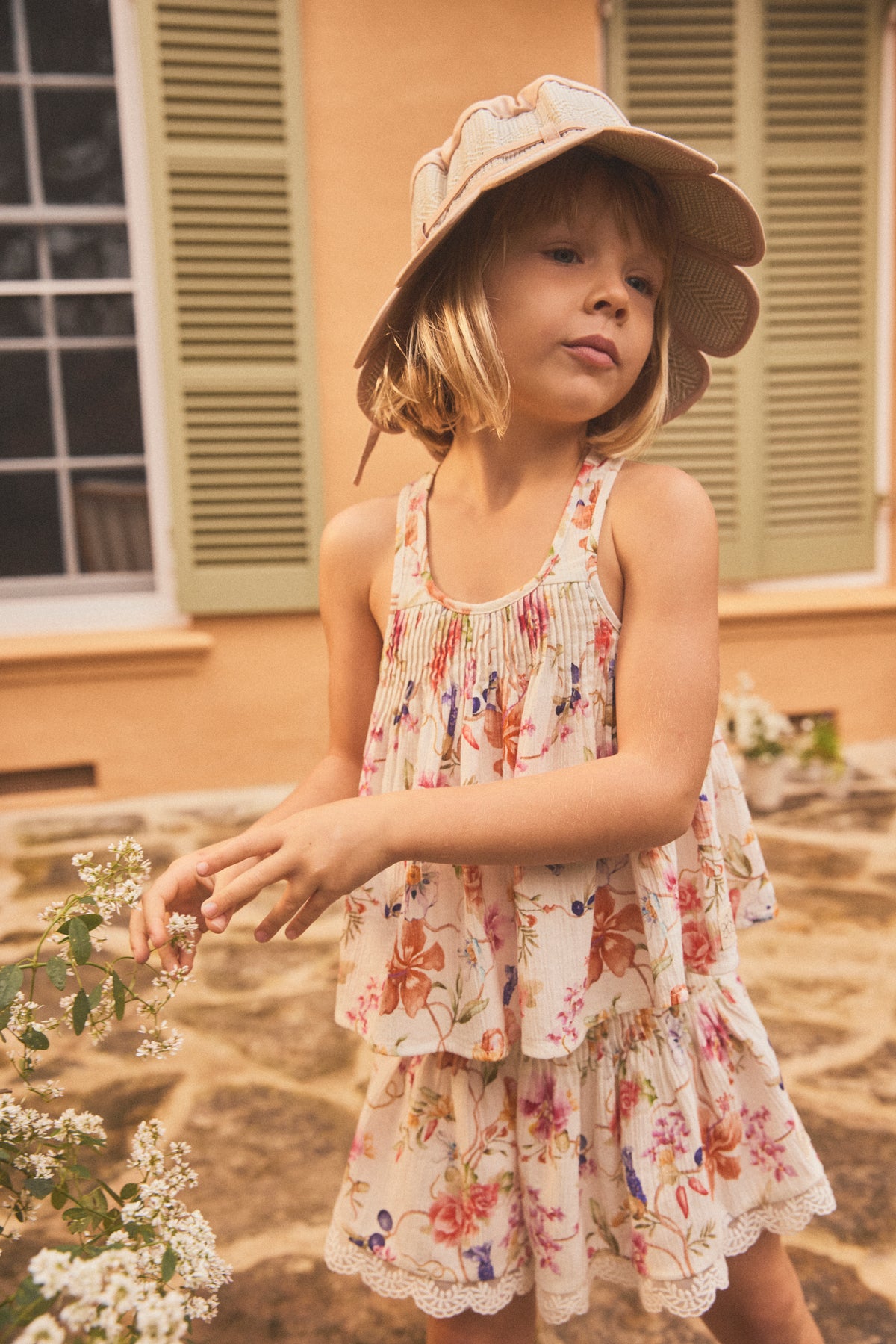 A young girl wearing a cami with a colorful floral print and a matching wide-brimmed sun hat, standing outdoors in front of a building with beige walls and green shutters.