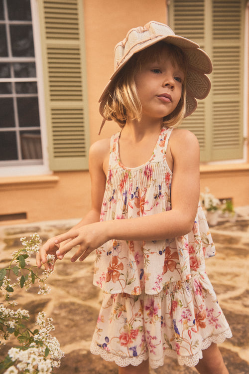 A young girl wearing a cami with a colorful floral print and a matching wide-brimmed sun hat, standing outdoors in front of a building with beige walls and green shutters.