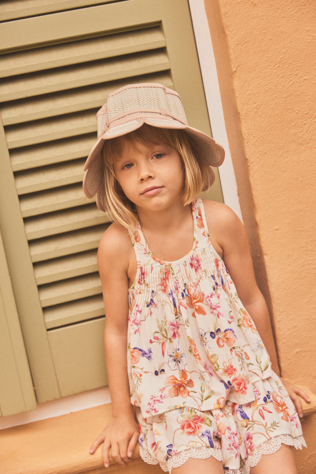 A young girl with blonde hair wearing a beige sun hat and a cream-colored floral matching set with a pleated cami, leaning against a warm orange wall next to a green shuttered window.
