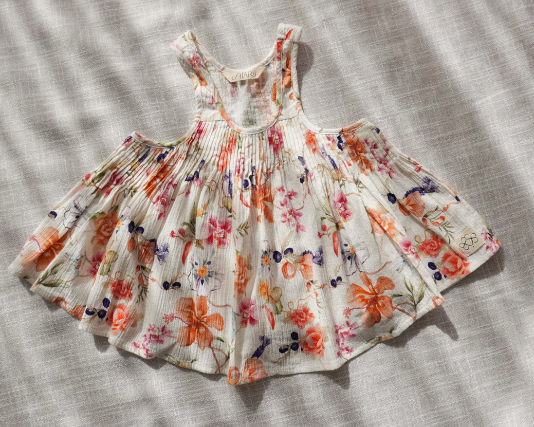 A sleeveless white cami with a colorful floral print and pleated details, laid flat on a textured white linen fabric background.