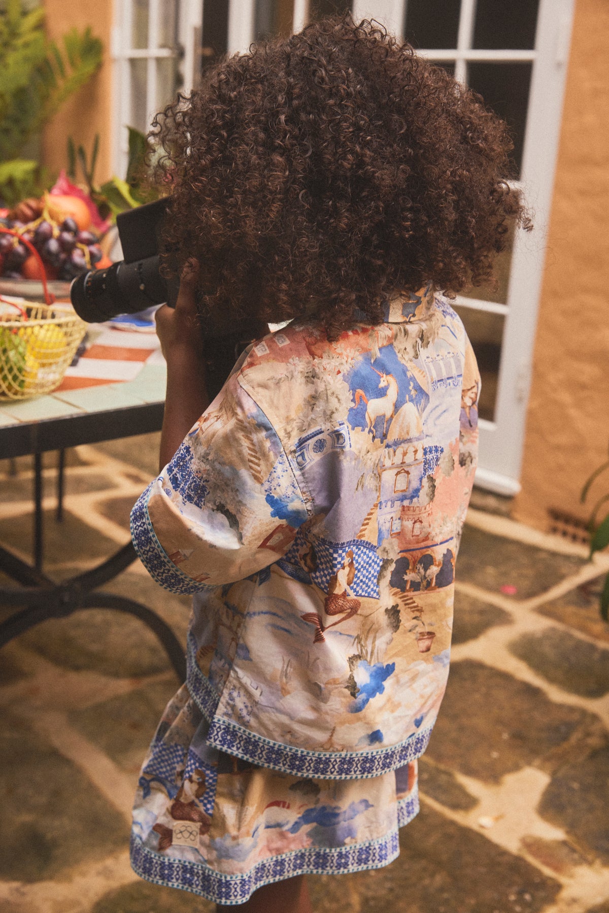 A child with curly dark hair seen from behind, wearing a short-sleeve shirt and short matching  set with a hand painted illustrated print and blue patterned trim, standing on a stone patio.