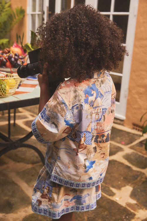 A child with curly dark hair seen from behind, wearing a short-sleeve shirt and short matching  set with a hand painted illustrated print and blue patterned trim, standing on a stone patio.