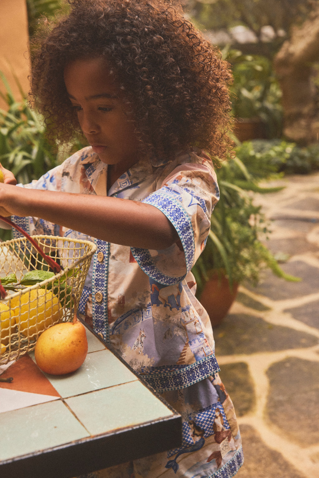A young girl with curly brown hair wearing a patterned short-sleeve shirt stands at a tiled table outdoors, reaching into a wire basket of fruit. The background shows a garden patio setting with plants and stone paving.