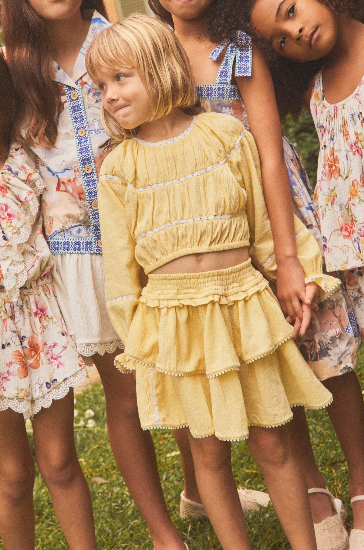 Five young girls wearing matching floral and pastel summer dresses pose together outdoors on a grassy lawn in front of a warm-toned house.