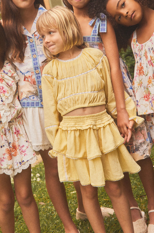Five young girls wearing matching floral and pastel summer dresses pose together outdoors on a grassy lawn in front of a warm-toned house.