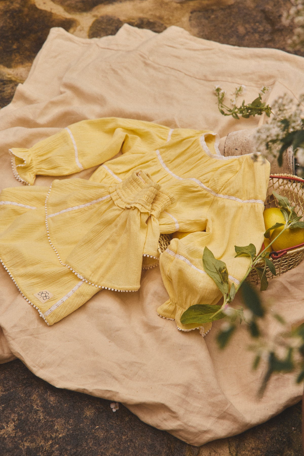 A soft yellow baby outfit set, including a long-sleeve blouse and matching skirt, styled on a beige linen cloth with a basket of lemons and greenery.