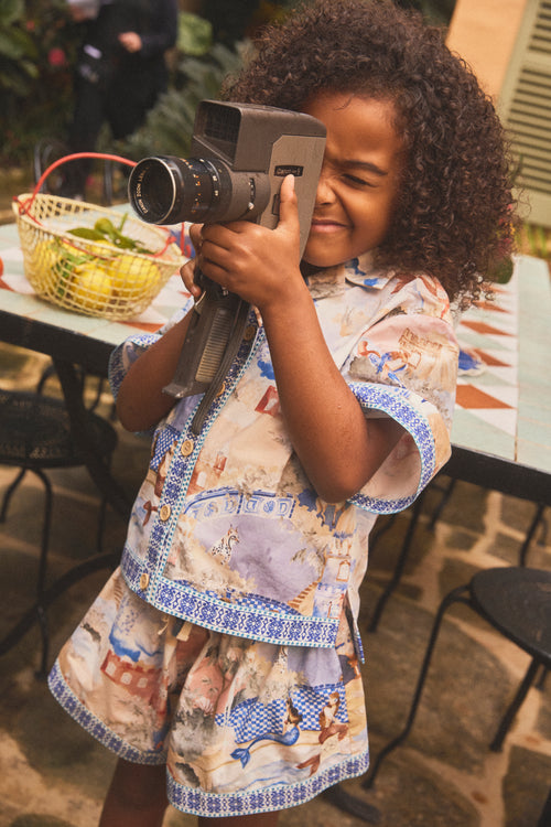 A young girl with curly dark hair holds a vintage-style camera up to their face, wearing a matching set with a button down shirt and shorts in a hand illustrated print and blue patterned trim, standing in an outdoor patio setting.