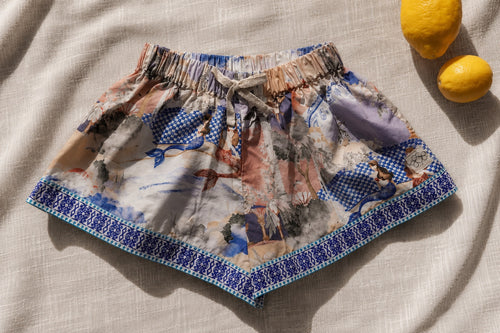 A pair of girl's shorts featuring a hand illustrated print with blue tiles borders and a drawstring waist, styled flat-lay on a beige linen fabric with two lemons in the corner.