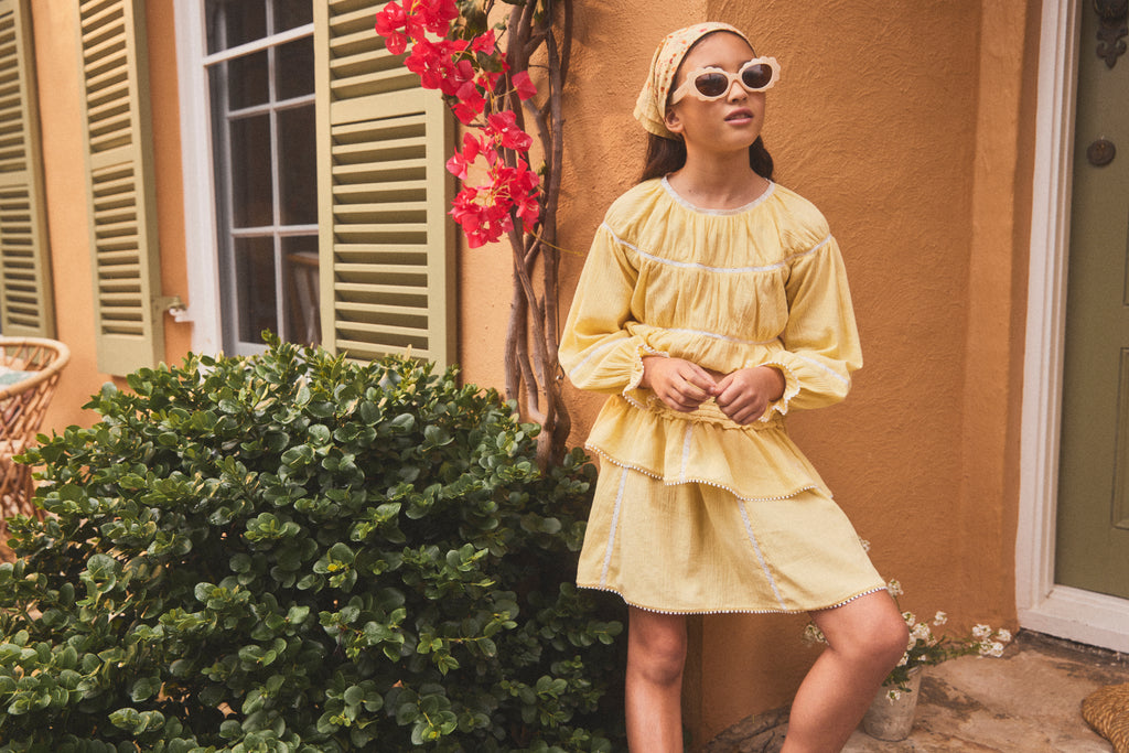 A young girl wearing a light yellow ruffle skirt, a matching yellow headscarf, and white sunglasses stands outdoors in front of a warm ochre-colored stucco wall with green shutters and a bush.