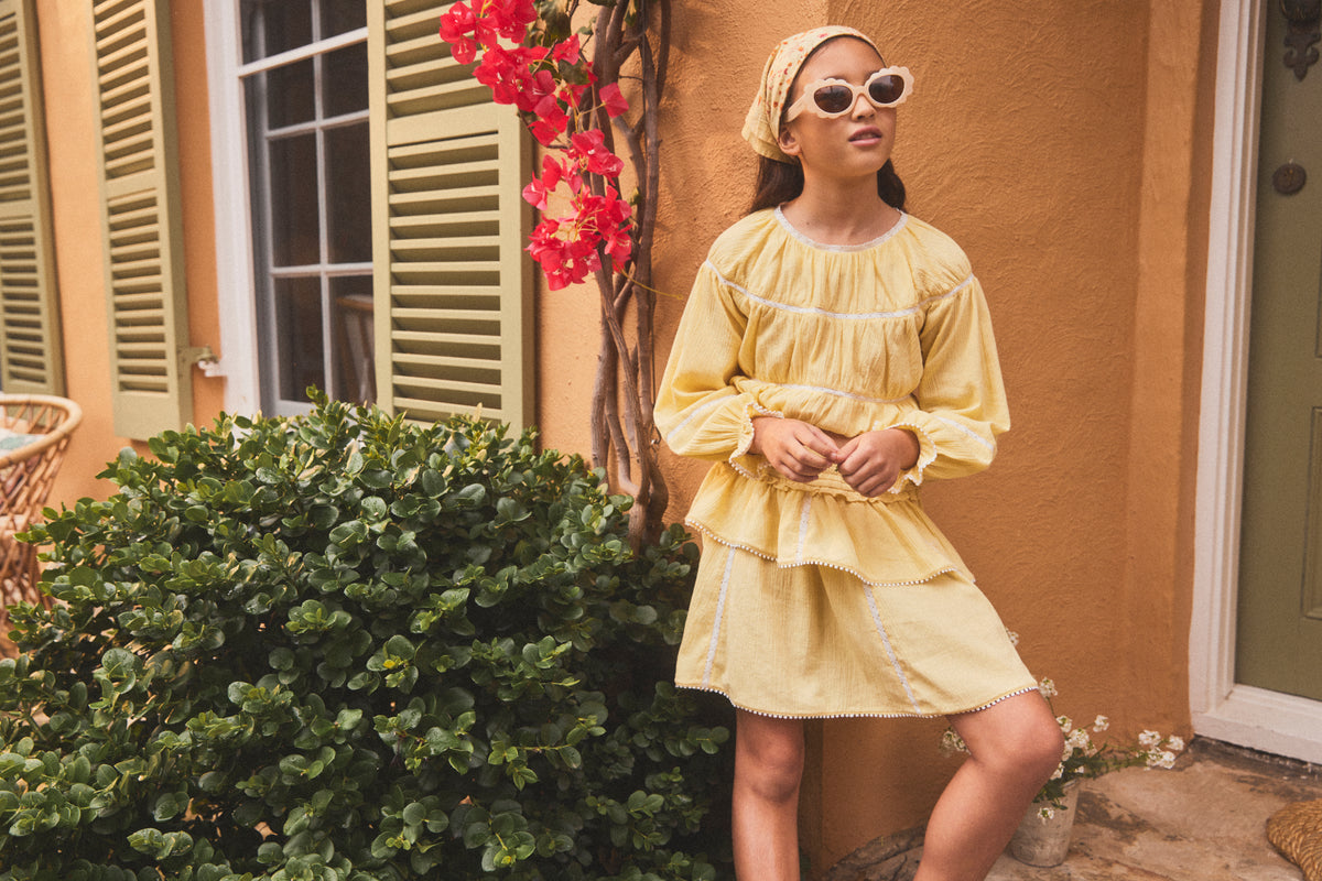A young girl wearing a light yellow ruffle skirt, a matching yellow headscarf, and white sunglasses stands outdoors in front of a warm ochre-colored stucco wall with green shutters and a bush.