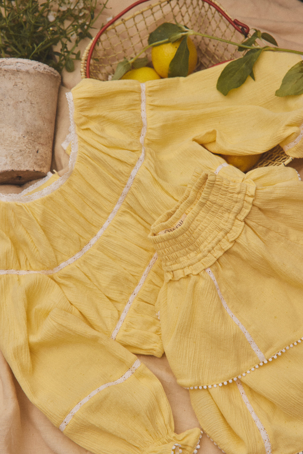 A yellow cotton matching set with white embroidered lines and ruffled trim, styled in a flat lay with lemons, a basket, and a potted plant.