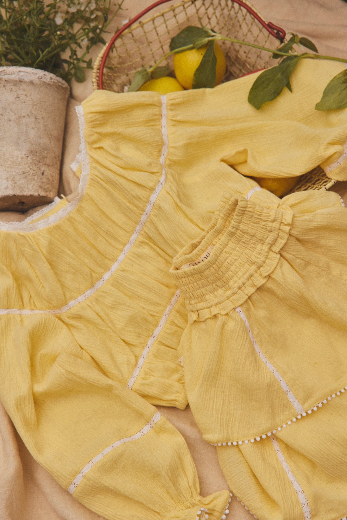 A yellow cotton matching set with white embroidered lines and ruffled trim, styled in a flat lay with lemons, a basket, and a potted plant.