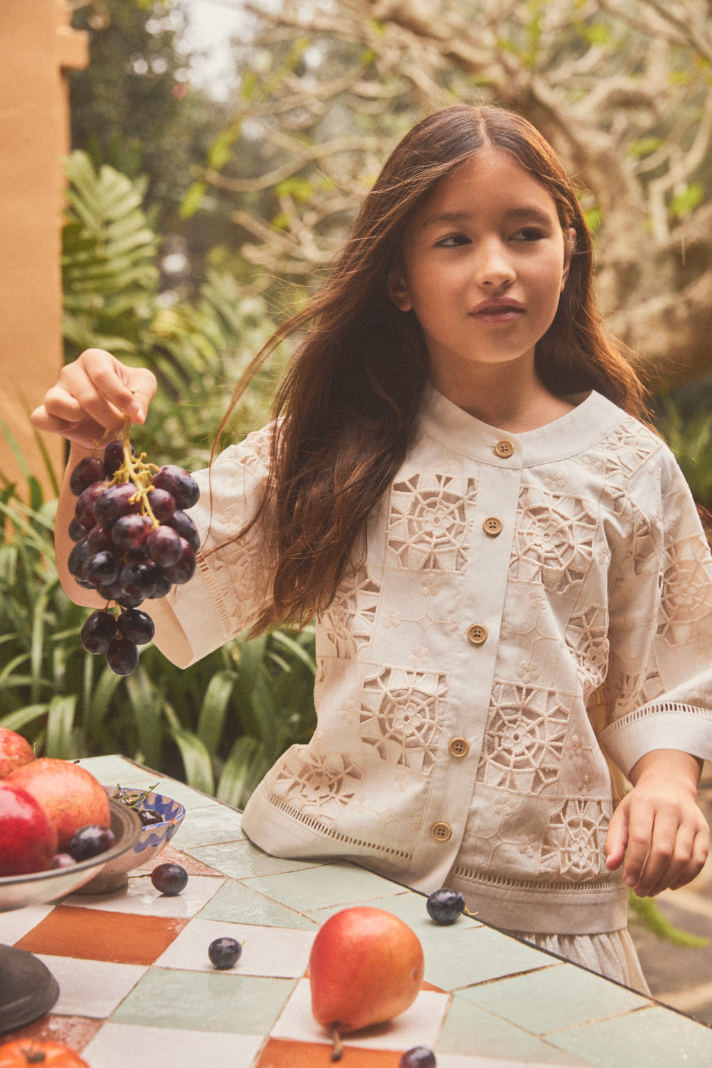 A young girl with long brown hair stands outdoors at a tiled table, wearing a cream-colored lace button down shirt with wooden buttons. She holds a bunch of dark purple grapes in one hand, with apples and grapes on the table in front of her.