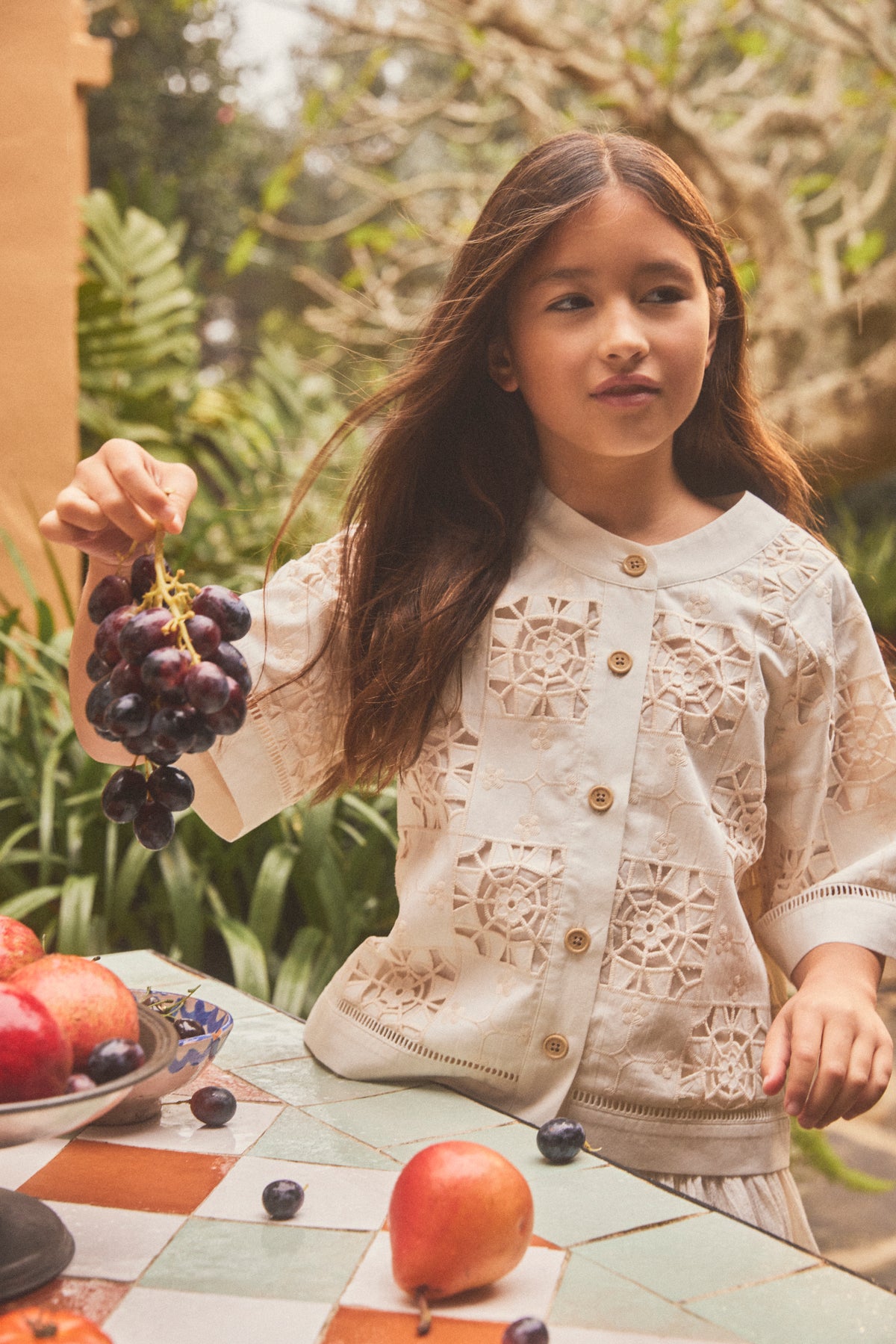A young girl with long brown hair stands outdoors at a tiled table, wearing a cream-colored lace button down shirt with wooden buttons. She holds a bunch of dark purple grapes in one hand, with apples and grapes on the table in front of her.