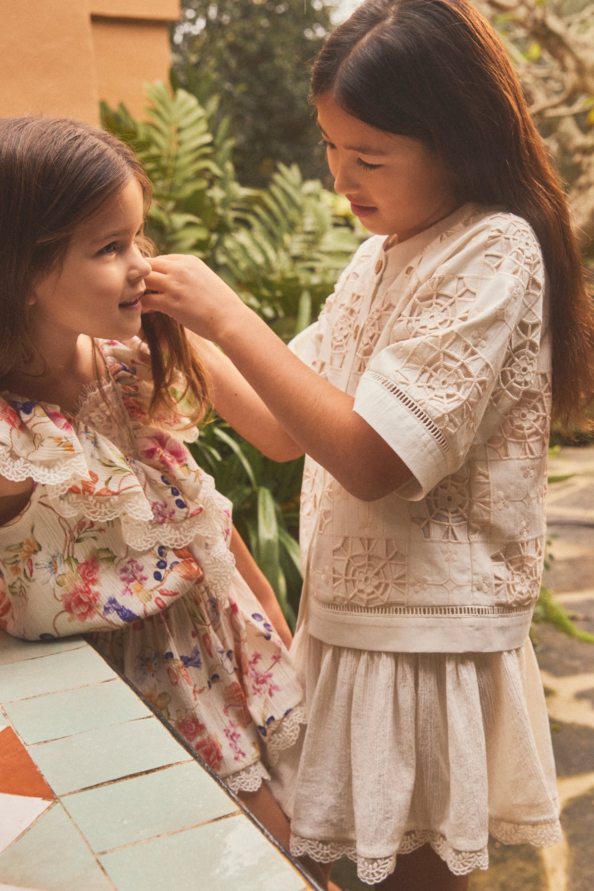 Two young girls lace and printed dresses in a warm, outdoor setting. The girl on the right wears a cream-colored lace button down shirt with floral lace details, while the girl on the left wears a matching floral print dress.