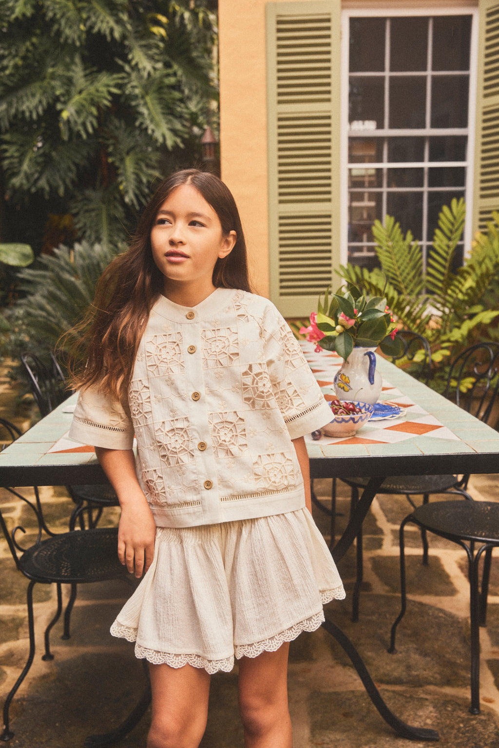 A young girl stands outdoors in a garden courtyard wearing a cream-colored two-piece outfit consisting of a short-sleeve button-front lace top and a matching flared short with lace trims.