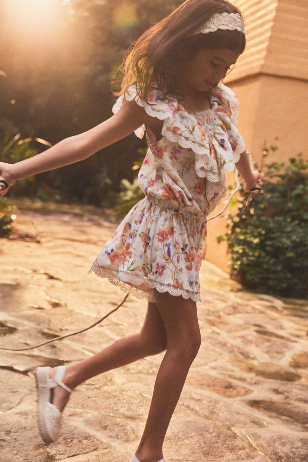 A young girl wearing a white ruffled floral playsuit and white shoes is captured mid-spin while playing outdoors on a stone path, with warm sunlight and soft-focus greenery in the background.