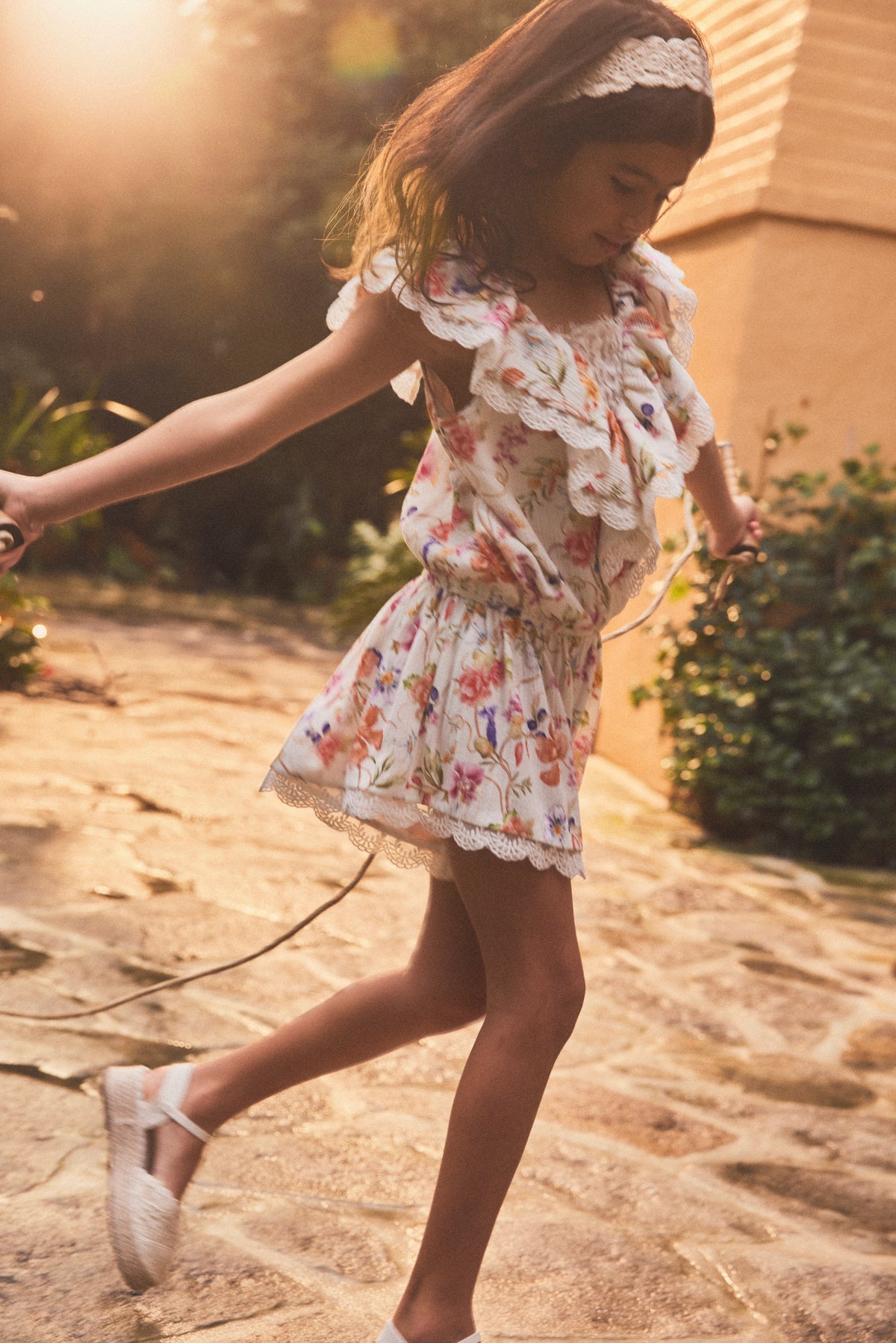 A young girl wearing a white ruffled floral playsuit and white shoes is captured mid-spin while playing outdoors on a stone path, with warm sunlight and soft-focus greenery in the background.