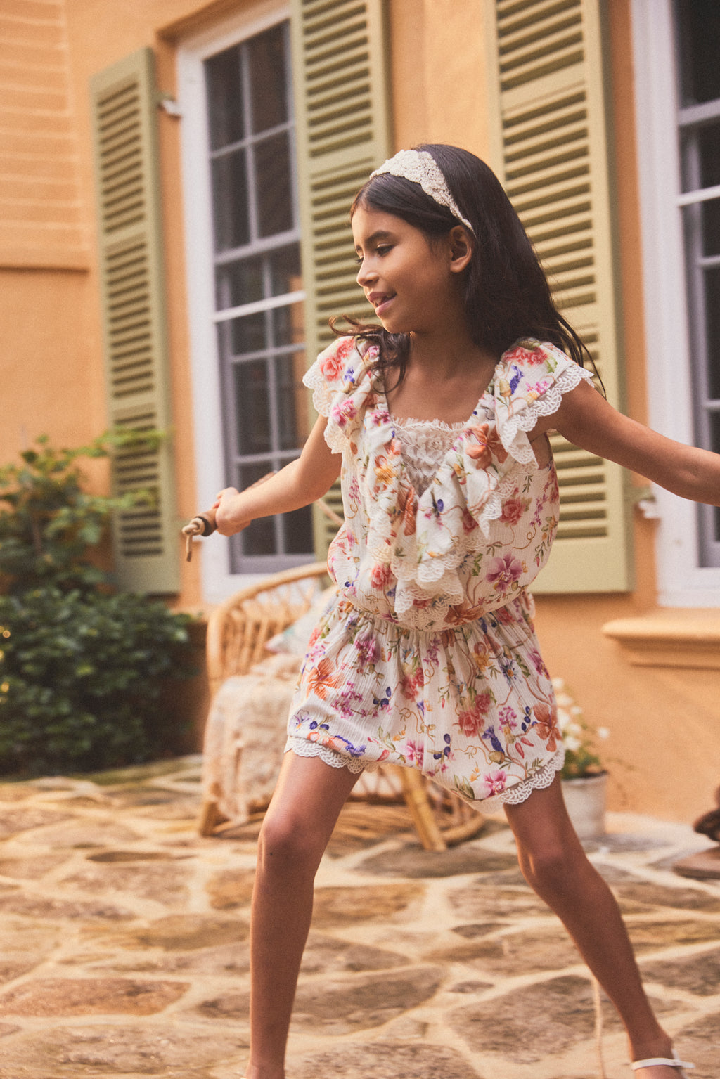 A young girl wearing a floral print ruffle dress and a matching headband is captured mid-dance in an outdoor courtyard setting with stone flooring and a warm yellow building in the background.