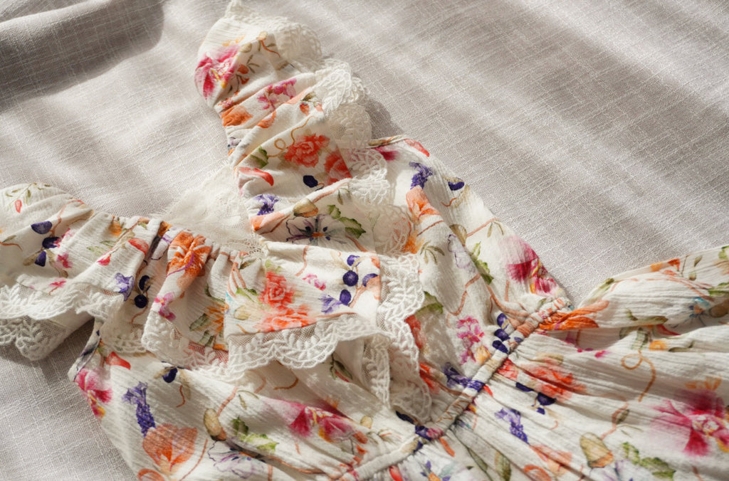 A close-up of a white playsuit with a colorful floral print and lace trim, laid out on a textured light fabric background.