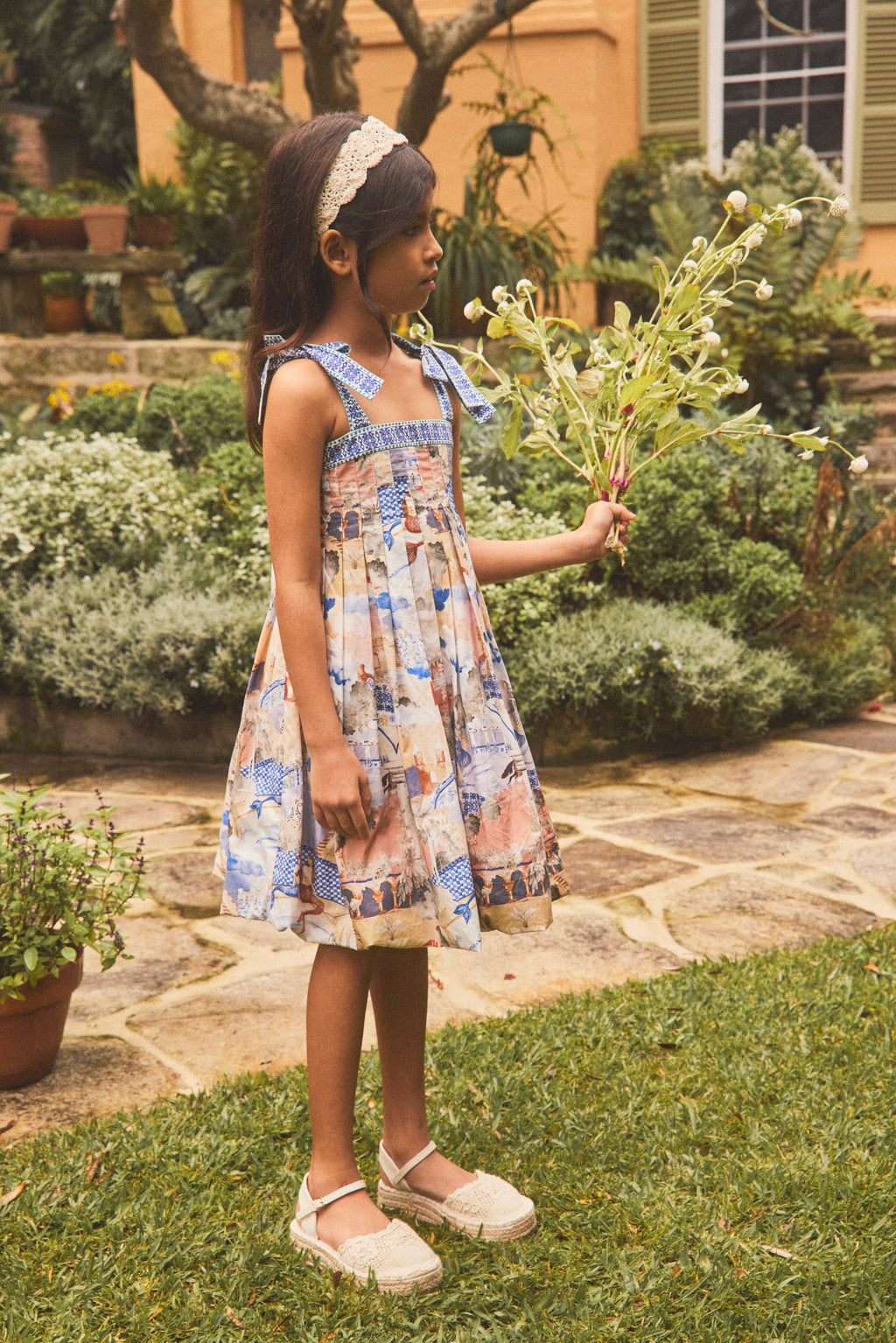 A young girl stands on a stone path in a garden wearing a sleeveless dress with a hand painted print, holding a bouquet of white flowers.