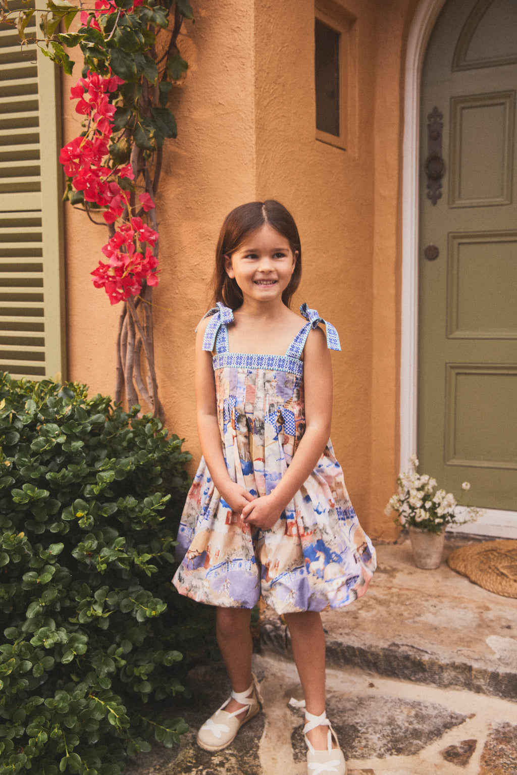 A young girl stands on stone steps in front of a stucco building, wearing a sleeveless dress with a colorful hand painted illustrated print and white shoes.