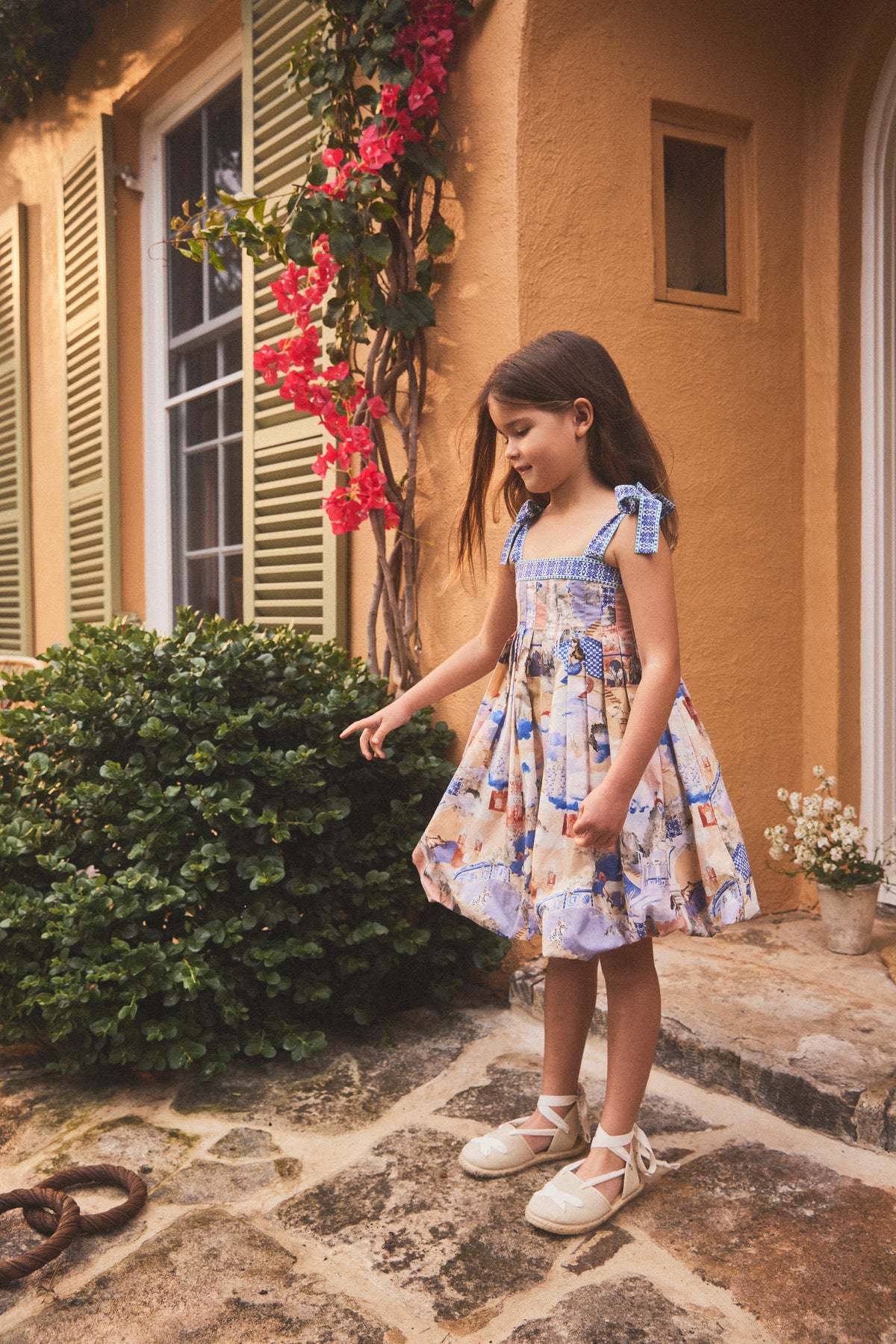 A young girl wearing a colorful patterned sundress and white lace-up sandals stands on a stone patio in front of a warm yellow stucco building with green shutters and red flowers.