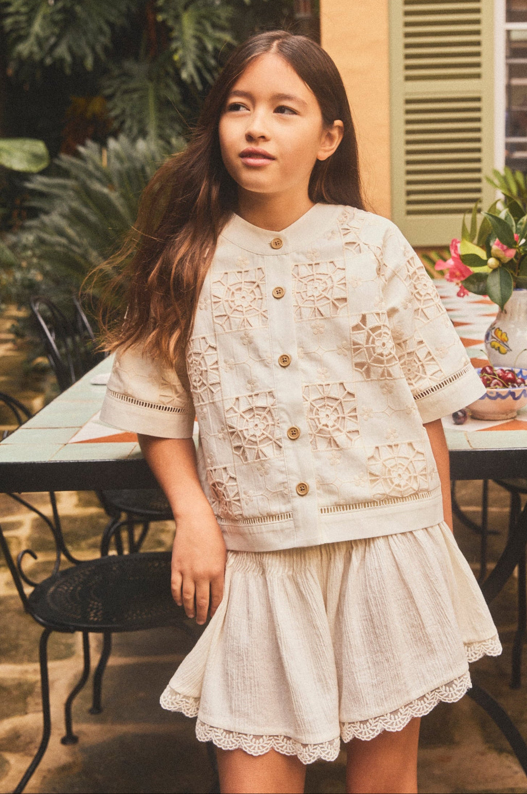A young girl stands outdoors in front of a table and house wall, wearing a cream-colored short-sleeve button-front top and matching flared skirt with lace trim.