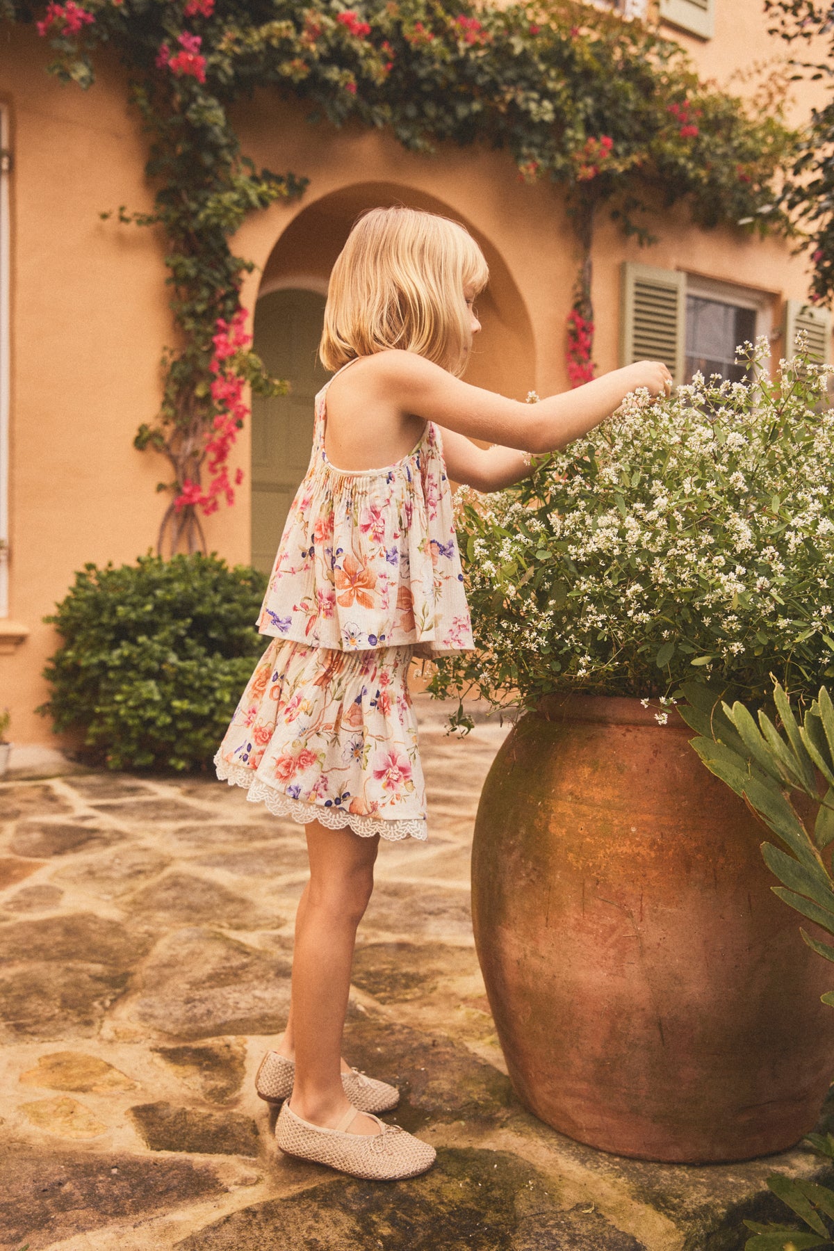 A young blonde girl in a sleeveless floral matching set with top and shorts stands on a stone patio beside a large copper planter filled with white flowers, with a warm Mediterranean-style building in the background.