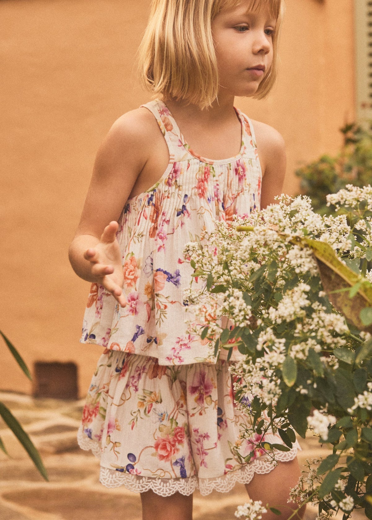 A young girl with blonde hair wearing a sleeveless tiered dress with a colorful floral print, standing outdoors next to a large bush of white flowers.