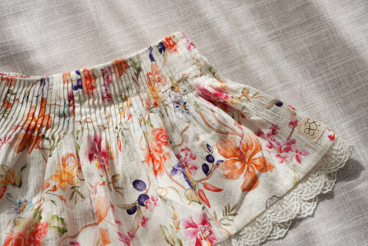 A close-up of a white cotton short with a colorful floral print, featuring a  pleated waistband and a lace hem.