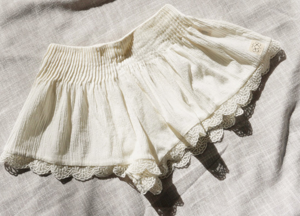 Cream crinkle cotton shorts with a lace hem, laid flat on a textured linen fabric background.