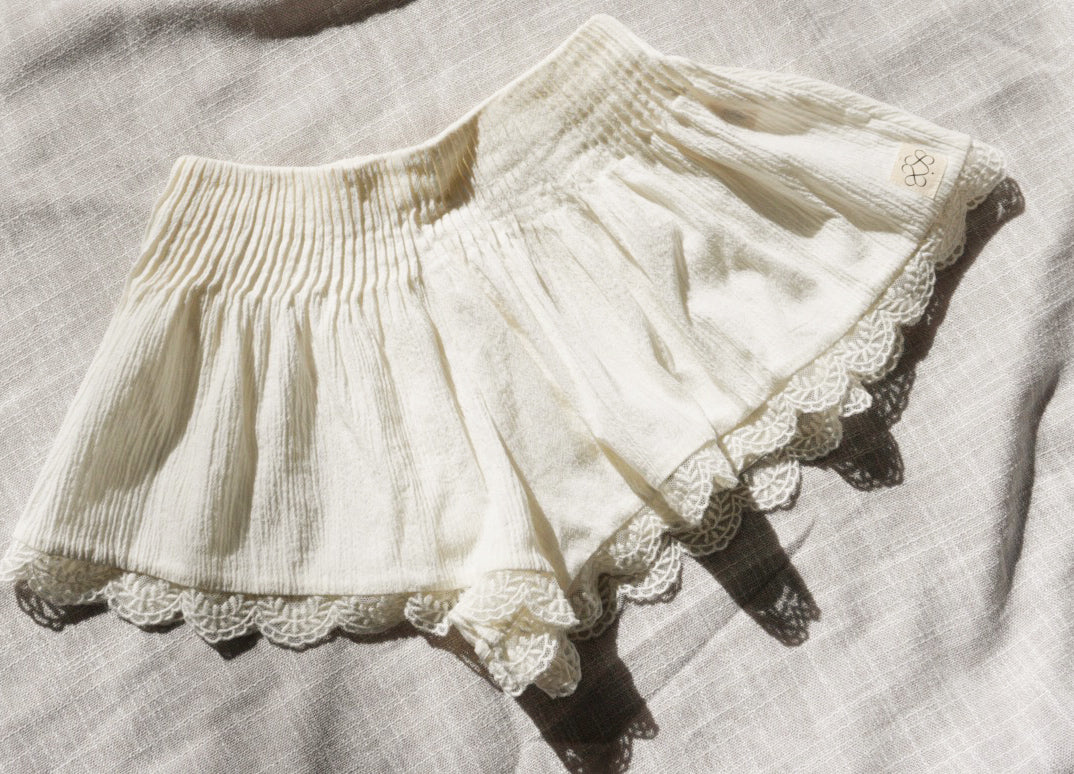 Cream crinkle cotton shorts with a lace hem, laid flat on a textured linen fabric background.