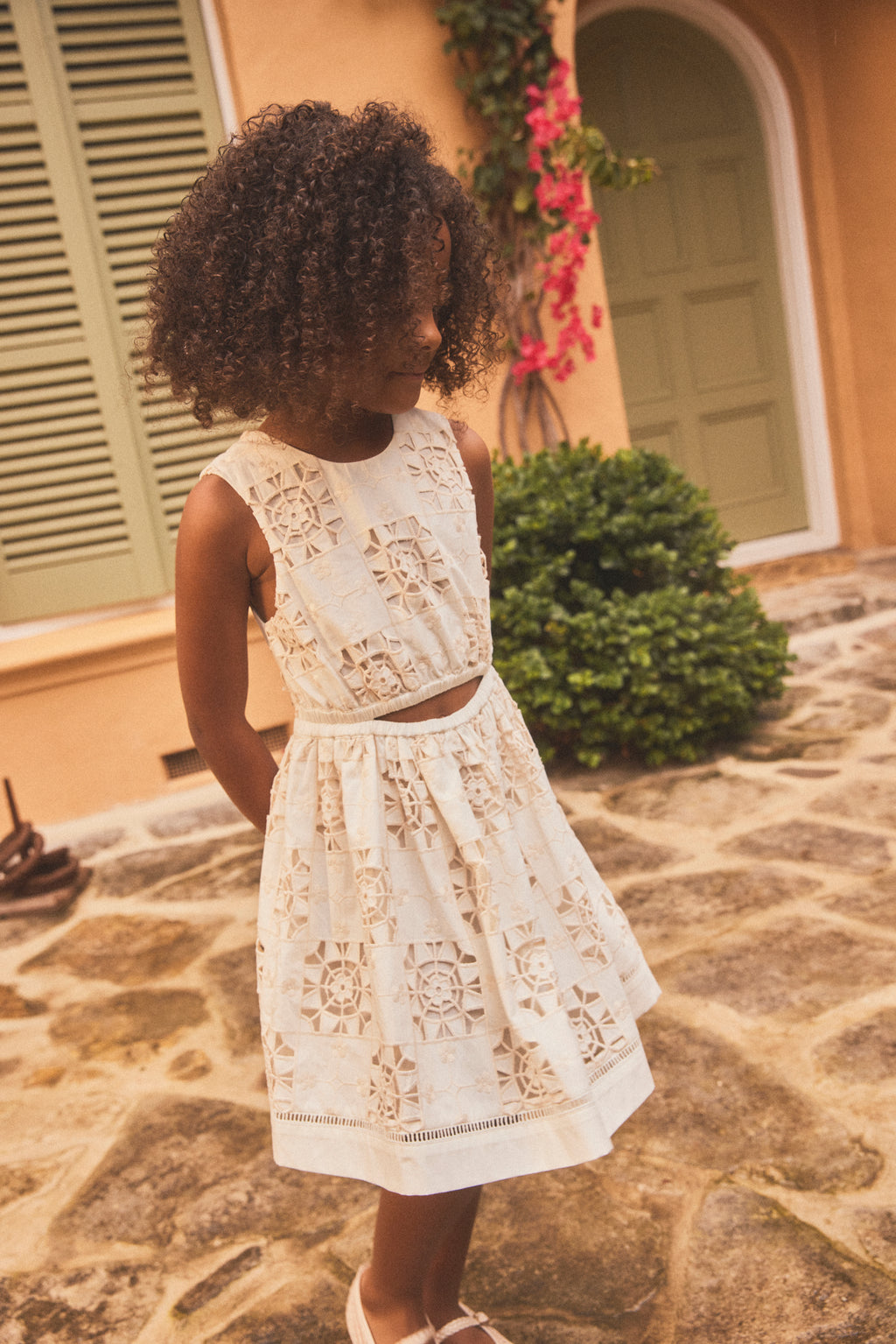 A young girl with curly dark hair wearing a sleeveless white lace dress , standing outdoors on a stone patio in front of a building with green shutters and a door.