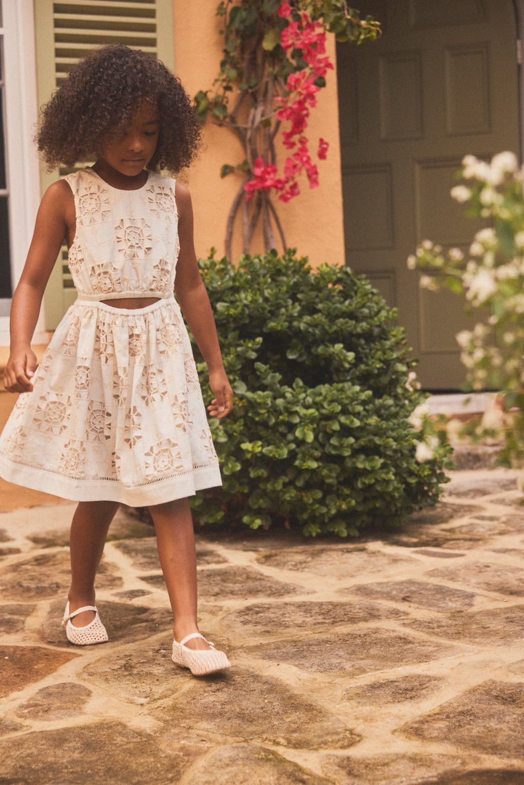 A young girl with curly dark hair wearing a sleeveless cream lace dress with a matching skirt and white Mary Jane shoes, walking on a stone patio in front of a house with greenery.