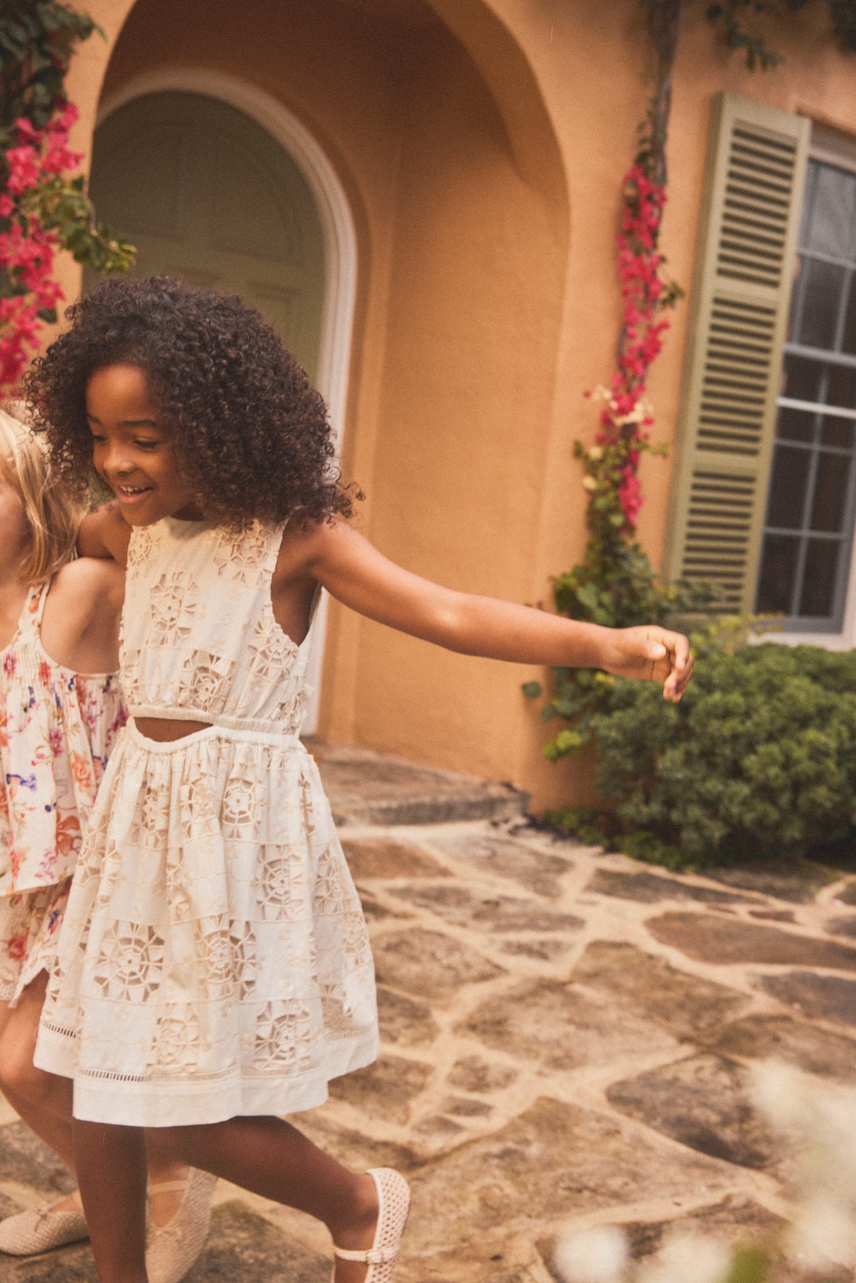 A young girl with curly dark hair wearing a sleeveless cream lace dress walks outdoors on a stone patio in front of a warm orange building with green shutters and pink flowers.