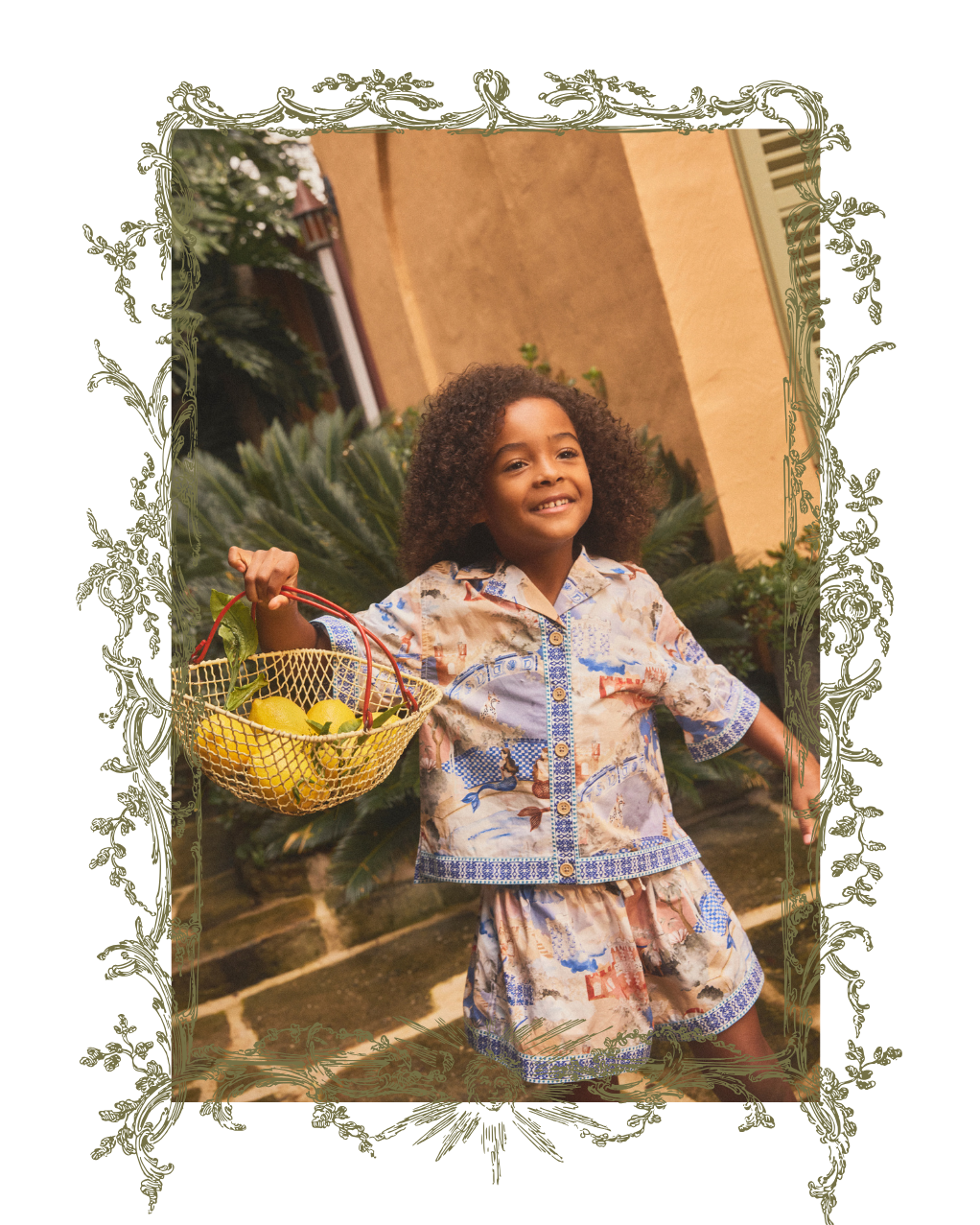A smiling child wearing a matching printed pajama set stands outdoors holding a woven basket of fruit. The pajamas have a light cream base with blue, red, and tan illustrated motifs.